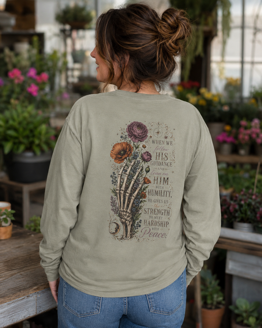 A woman viewed from behind wearing a sage green long-sleeve graphic sweatshirt with a detailed back print featuring a skeleton hand, flowers, and inspirational text, set in a plant-filled indoor environment.
