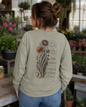 A woman viewed from behind wearing a sage green long-sleeve graphic sweatshirt with a detailed back print featuring a skeleton hand, flowers, and inspirational text, set in a plant-filled indoor environment.