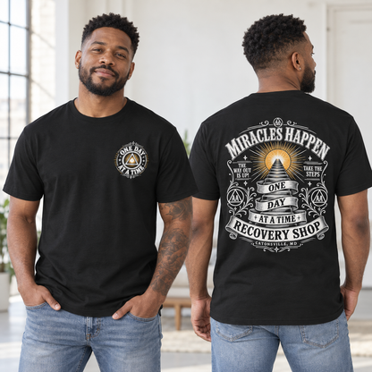 A man modeling a black graphic T-shirt, shown from both the front and back. The front features a small circular chest graphic reading 'ONE DAY AT A TIME' with a pyramid and triangle.