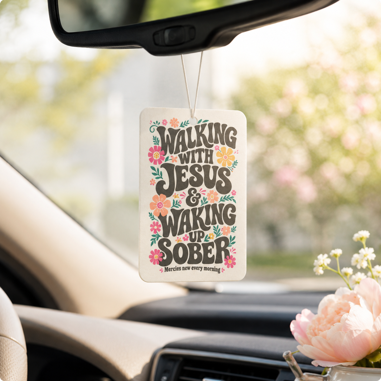 A rectangular car air freshener hanging from a rearview mirror inside a car. The white card has the phrase “WALKING WITH JESUS & WAKING UP SOBER” in dark lettering, surrounded by pink, orange, and yellow flowers with green leaves.