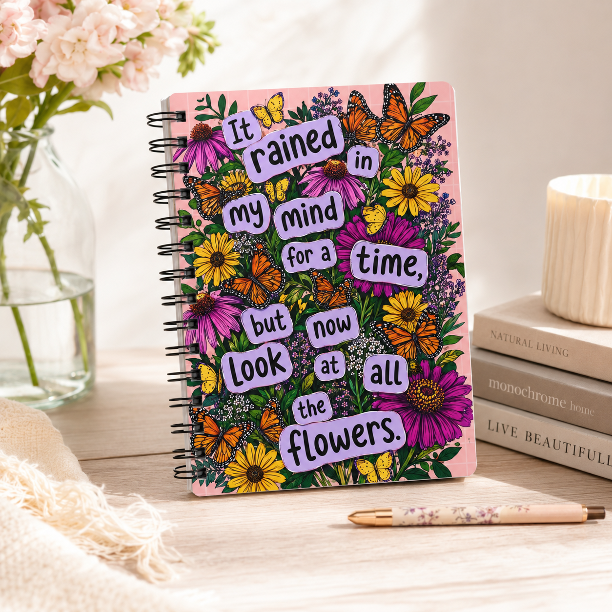 A pink spiral-bound notebook with a floral cover design and motivational text, styled on a wooden tabletop with a pen, stacked books, a candle, and flowers in the background.