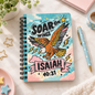 A spiral-bound notebook with a colorful illustrated cover featuring an eagle, clouds, stars, and the text 'Soar on Wings Isaiah 40:31,' styled on a light marble surface with a pen, plant, and pastel props.
