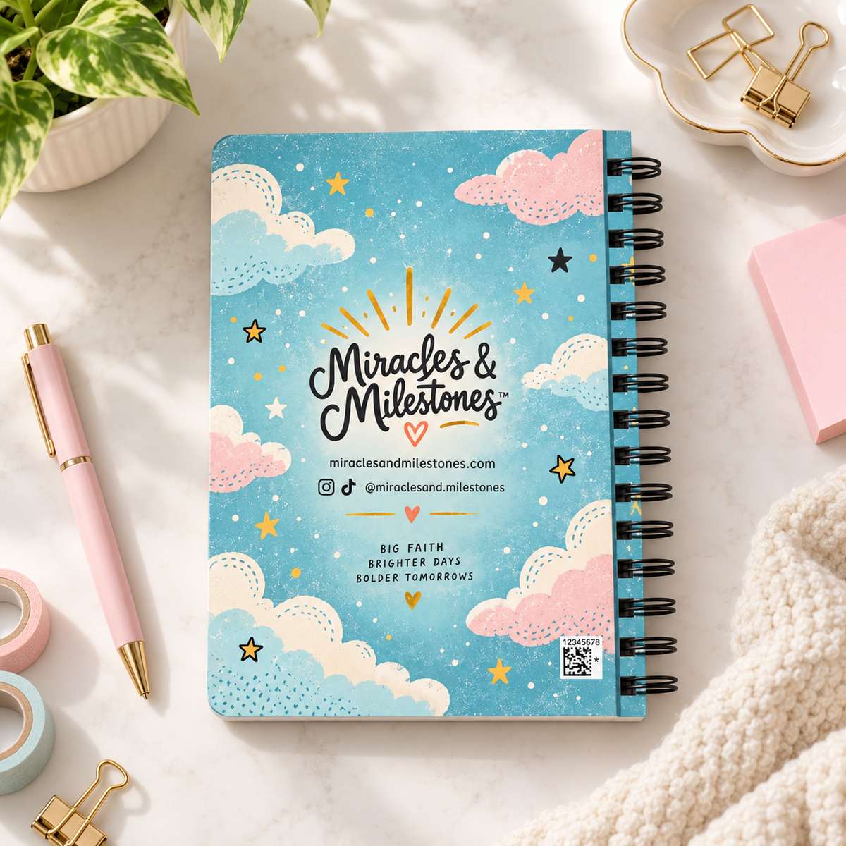 A spiral-bound notebook with a blue cover decorated with pink and cream clouds, white stars, and gold sun rays. The cover features the text 'Miracles & Milestones' in a decorative script, along with social media handles and website information.