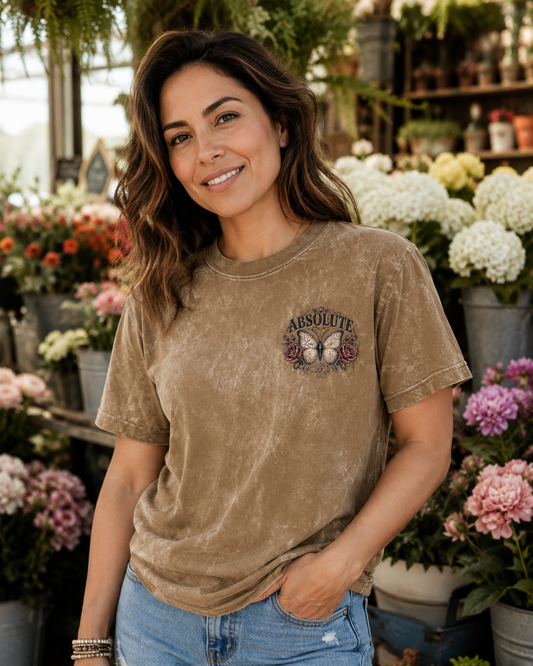 A woman wearing a brown mineral-wash t-shirt with a butterfly graphic and the text 'ABSOLUTE' on the chest, standing in a flower shop.