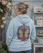 A woman wearing a light blue hoodie, viewed from behind, featuring a large graphic print of a butterfly surrounded by roses and the word 'ABSOLUTELY' on the back. She is standing in a garden setting with flowers and rustic furniture in the background.