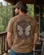 A man wearing a brown t-shirt with a large butterfly and rose graphic on the back, standing on a rustic wooden porch.