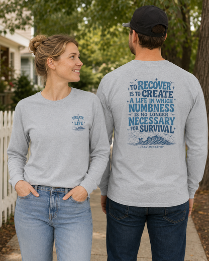 A man and a woman standing outdoors on a sidewalk, both wearing light heather gray long-sleeve t-shirts with blue text and graphic prints. The woman's shirt has a small chest print reading 'CREATE LIFE', and the man's shirt has a large back print with a quote by Jean McKeown.
