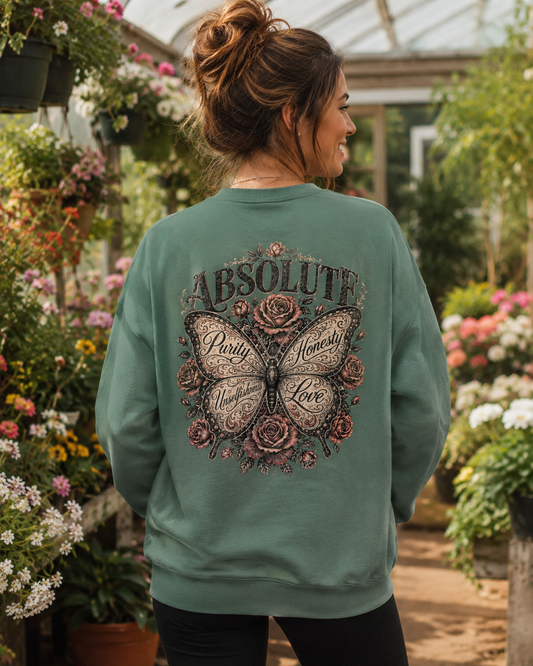 A woman viewed from behind wearing a sage green crewneck sweatshirt with a large butterfly and floral graphic on the back that reads 'ABSOLUTE Party & Nectar Love'. The setting is a lush, sunlit greenhouse filled with flowers.
