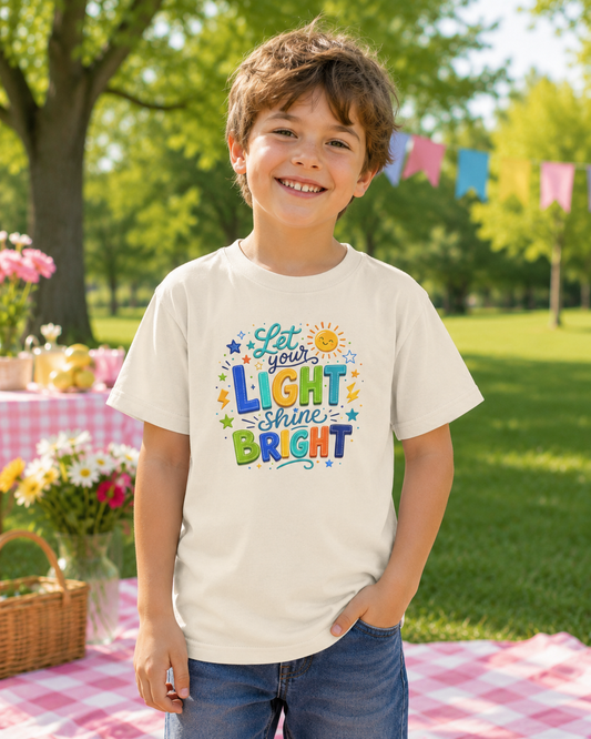 A young boy smiling outdoors on a picnic blanket, wearing a cream-colored t-shirt with a colorful graphic that reads 'Let your LIGHT shine BRIGHT' surrounded by suns and stars, paired with blue jeans.