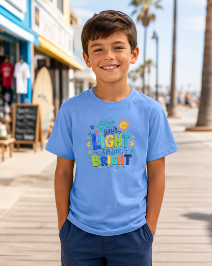 A smiling young boy wearing a light blue graphic t-shirt with the colorful text 'Let your LIGHT shine BRIGHT' and navy blue pants, standing on a sunny boardwalk.