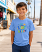 A smiling young boy wearing a light blue graphic t-shirt with the colorful text 'Let your LIGHT shine BRIGHT' and navy blue pants, standing on a sunny boardwalk.