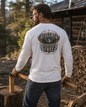 A man viewed from behind wearing a white long-sleeve t-shirt with a large back graphic that reads 'Redeemed Recovered Free' with wings and a cross. He is standing outdoors in front of a log cabin with wood stacked nearby.