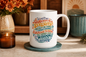 A white ceramic coffee mug with a colorful, decorative typographic religious quote printed on the side, sitting on a teal coaster on a tabletop with a lit candle and autumn flowers in the background.