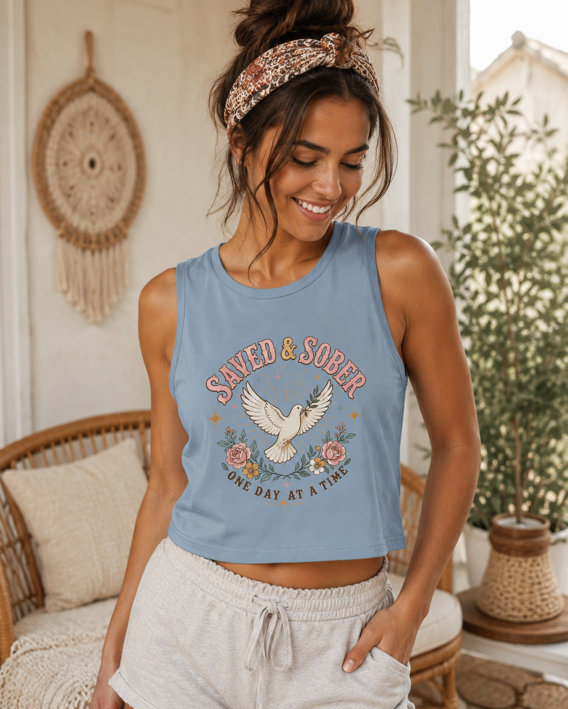 A woman wearing a light blue sleeveless crop top with a 'Saved & Sober' graphic featuring a dove and flowers, paired with light gray drawstring shorts in a cozy indoor setting.