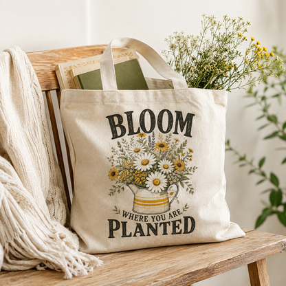 A natural canvas tote bag with the text 'BLOOM WHERE YOU ARE PLANTED' and a floral illustration, resting on a wooden bench with a cream knit blanket and dried flowers in a cozy indoor setting.