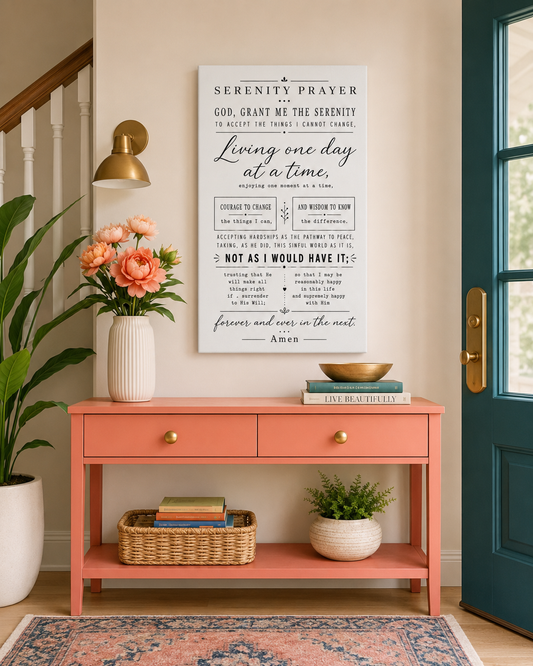 A framed 'Serenity Prayer' wall art print hangs on a beige wall in a styled entryway above a coral console table, with flowers, a lamp, plants, and a teal door visible.