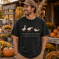 A man wearing a black graphic t-shirt with a white duck illustration and the text 'WALKING BY FAITH tripping constantly' stands in front of a rustic autumn-themed shop background with pumpkins and mums.