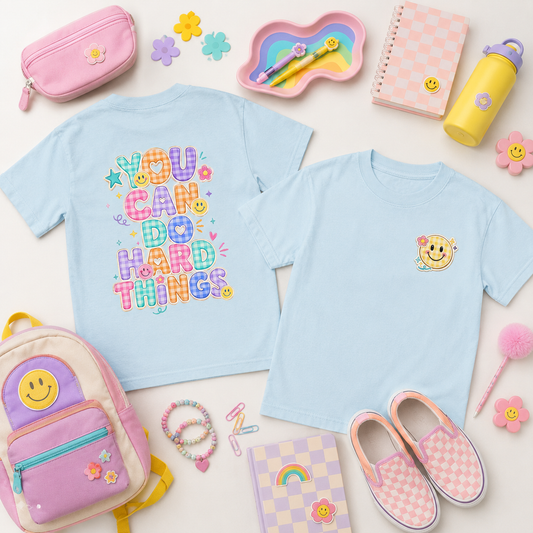 A flat-lay collection of pastel-colored school and play accessories, featuring two light blue t-shirts (one with a colorful 'YOU CAN DO HARD THINGS' graphic on the back, and one with a smiley face on the chest), a pink and purple backpack, a pink pencil case, pink-and-white checkered slip-on shoes, and other matching pastel items on a white background.